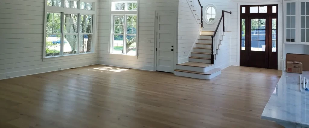 Bright living room with natural hardwood flooring, large windows, coffered ceiling, and open staircase with dark railing